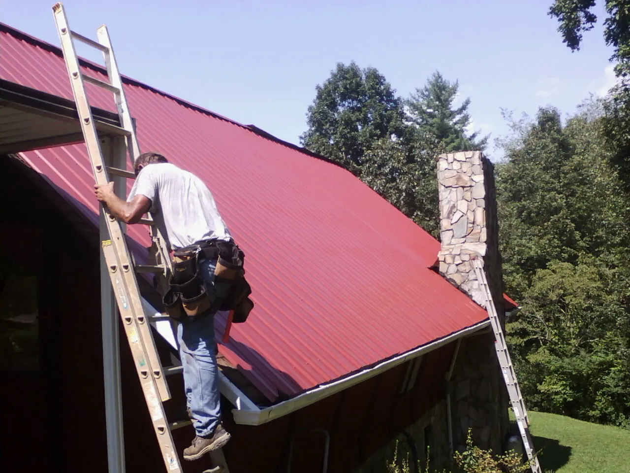 Roofer on a ladder beside a red metal roof and stone chimney — Soffit & Fascia Repair by Roofing Brothers in Cedarville